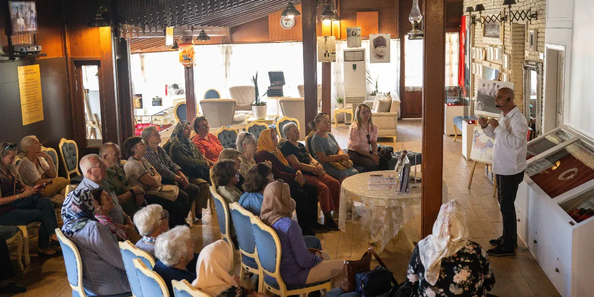 Ahşap ağırlıklı dekore edilmiş, tarihi bir atmosferi olan salonda, beyaz gömlekli bir erkek ayakta durarak dinleyici grubuna sunum yapıyor. Çoğunluğu kadınlardan oluşan, farklı yaş gruplarındaki izleyiciler, ahşap sandalyelerde oturarak konuşmacıyı dikkatle dinliyor. Salonun duvarlarında Atatürk portresi, çeşitli tarihi fotoğraflar ve bilgilendirme panoları yer alıyor. Sağ tarafta, üzerinde Türk bayrağı bulunan camlı bir sergi ünitesi ve konuşmacının yanında bir şövale üzerinde tarihi bir fotoğraf bulunuyor. Ortadaki yuvarlak masanın üzerinde ise kitaplar ve broşürler yer alıyor.