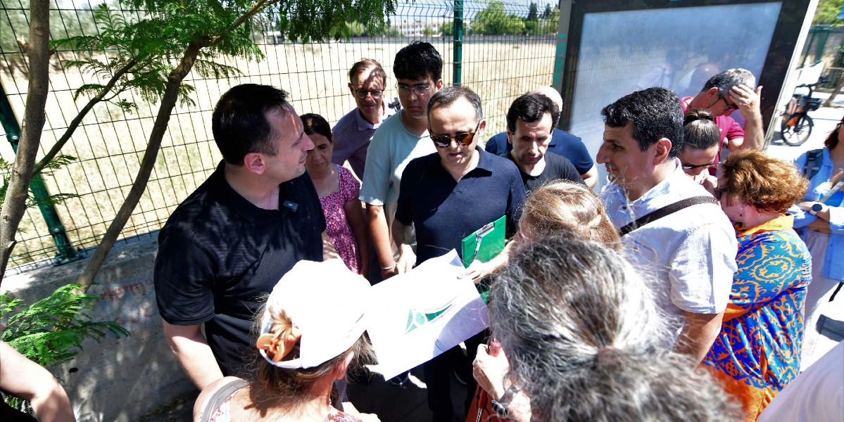 Açık havada, tel çitlerin önünde toplanmış bir grup insan, ellerindeki bir proje dosyasını veya haritayı inceleyerek fikir alışverişinde bulunuyor. Gruptaki kişiler, üzerinde Bornova Belediyesi logosu bulunan bir belgeyi dikkatle incelerken, aralarında engelli hakları ve erişilebilirlik üzerine bir değerlendirme yapıyor. Ortamda güneşli bir hava hakim ve katılımcılar, yerel yönetim temsilcileri ve vatandaşlardan oluşan karma bir grubu temsil ediyor.