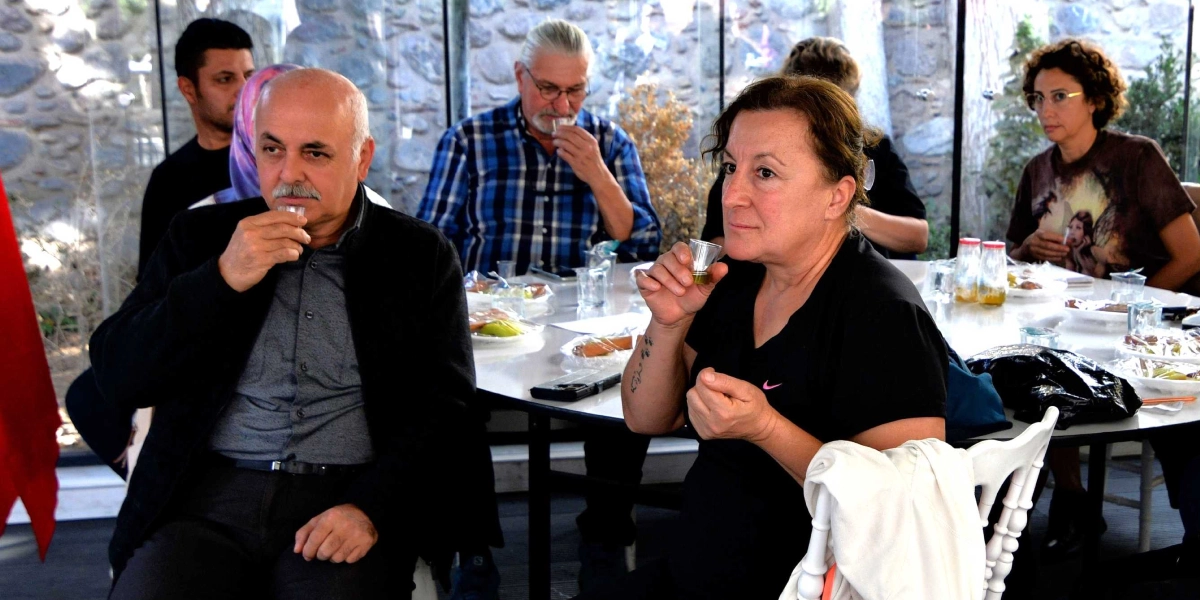 Bornova'daki zeytin hasadı etkinliğinde, bir masanın etrafında toplanmış bir grup insan küçük bardaklarda zeytinyağı tadımı yapıyor. Ön planda, siyah kıyafetli orta yaşlı bir erkek ve yanında siyah tişörtlü bir kadın ellerindeki küçük bardaklardan zeytinyağını tadarken görülüyor. Arka planda ise mavi ekose gömlekli bir erkek ve gözlüklü bir kadın da tadım yapmaya devam ediyor. Masanın üzerinde tadım için hazırlanmış küçük porsiyonlar ve zeytinyağı şişeleri bulunuyor.