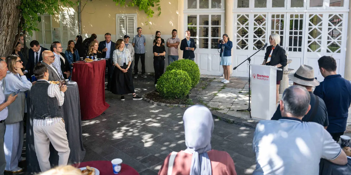 Tarihi bir binanın bahçesinde düzenlenen etkinlikte, üzerinde Bornova Belediyesi logosu bulunan kürsüde konuşma yapan kısa sarı saçlı bir kadın ve onu dinleyen katılımcı bir grup yer alıyor. Katılımcılar arasında ayakta duranlar ve kokteyl masalarının etrafında sohbet edenler bulunuyor. Bahçede yeşil çalılar, ağaçlar ve beyaz çerçeveli pencerelere sahip taş bir yapı görülüyor.