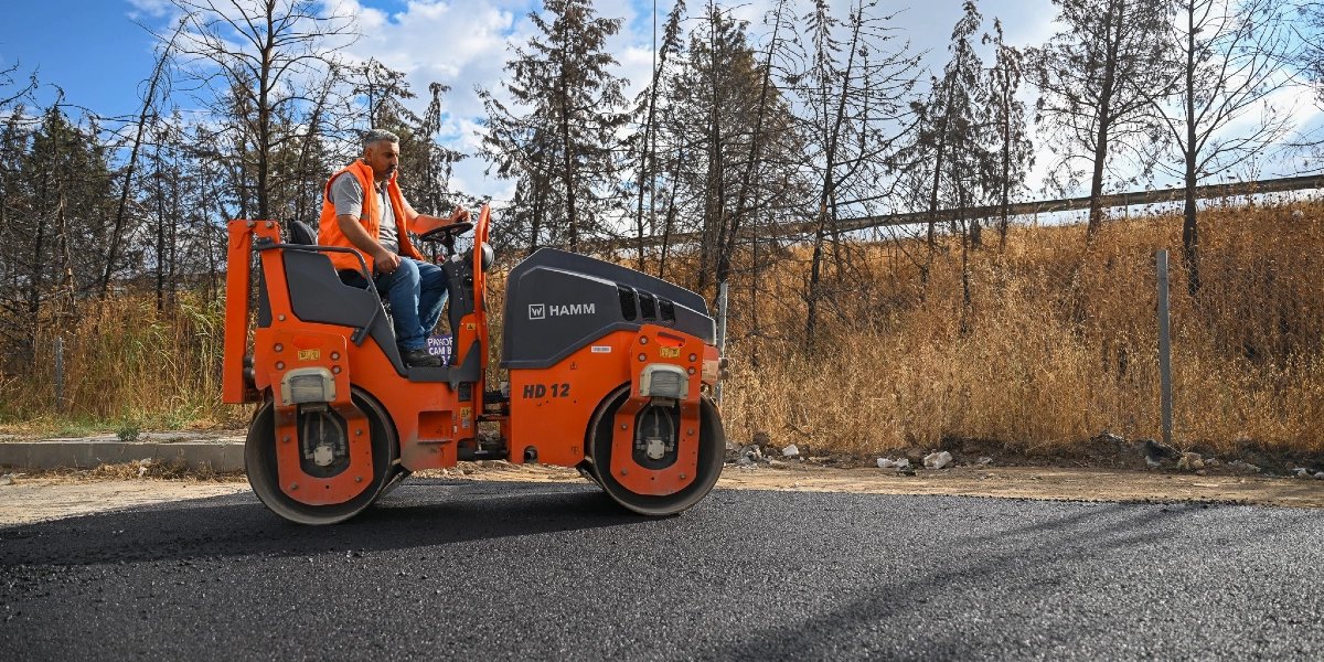 Turuncu yelekli bir operatör, Hamm HD 12 model silindir iş makinesini kullanarak yeni dökülmüş asfaltı düzleştiriyor. Arka planda kuru otlar ve ağaçların bulunduğu bir yol kenarı manzarası yer alıyor.