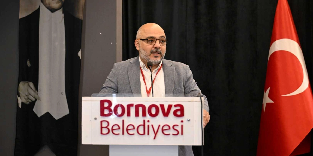 Gri ceketli, gözlüklü ve sakallı bir erkek, üzerinde Bornova Belediyesi yazılı bir kürsüde konuşma yapıyor. Arka planda sol tarafta Atatürk'ün fraklı bir portresi, sağ tarafta ise Türk bayrağı yer alıyor.