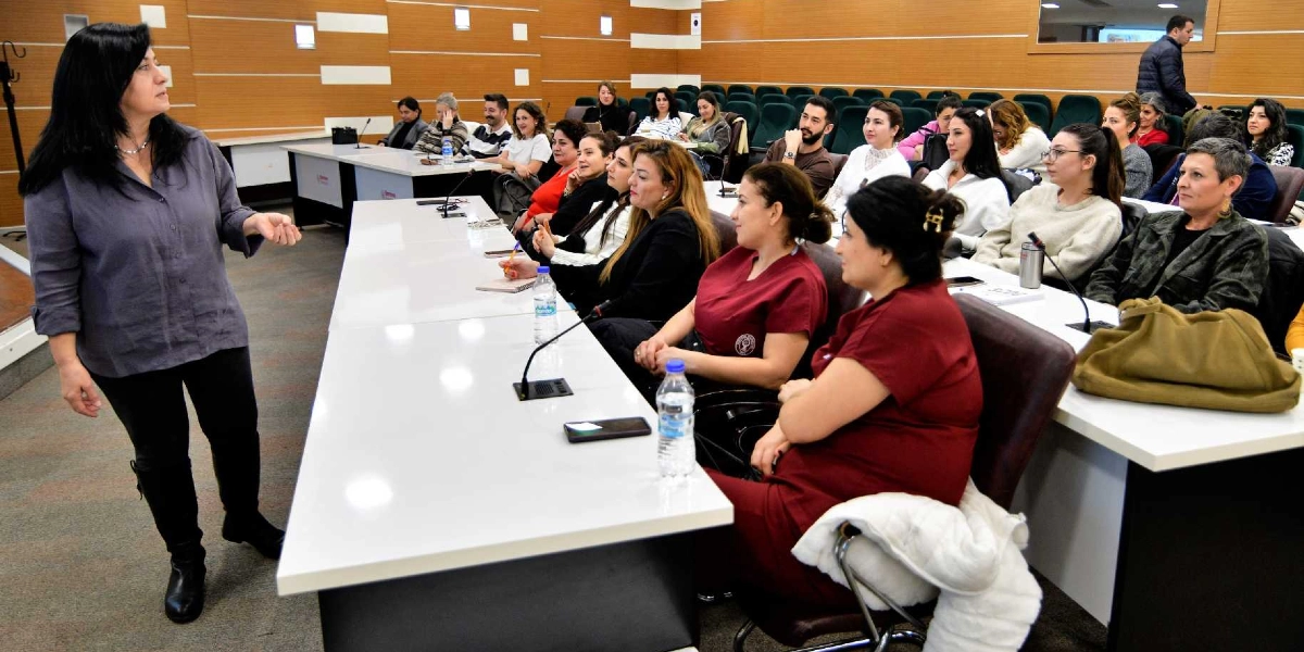 Bornova Belediyesi'nde düzenlenen bir toplantıda, gri gömlekli bir kadın katılımcılara hitap ederken, uzun beyaz masalar etrafında oturan çoğunluğu kadınlardan oluşan bir grup dinleyici onu dikkatle takip ediyor.