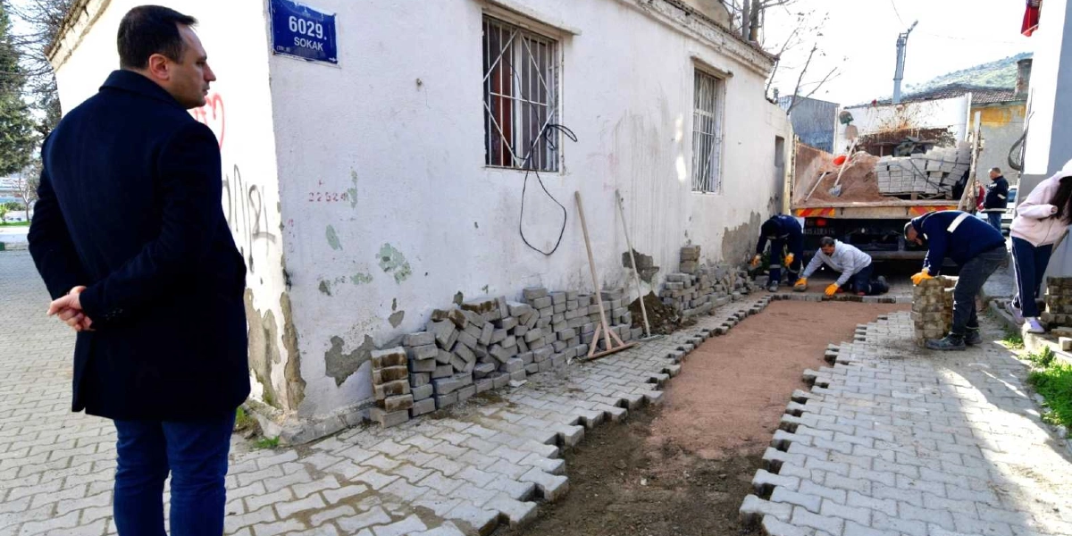 Koyu renkli ceket giymiş bir erkek, Bornova 6029. Sokak tabelasının bulunduğu beyaz bir binanın önünde devam eden yol yenileme çalışmalarını izliyor. İşçiler, bir kamyonun yanında kilit parke taşlarını döşeyerek kaldırım düzenlemesi yapıyor.