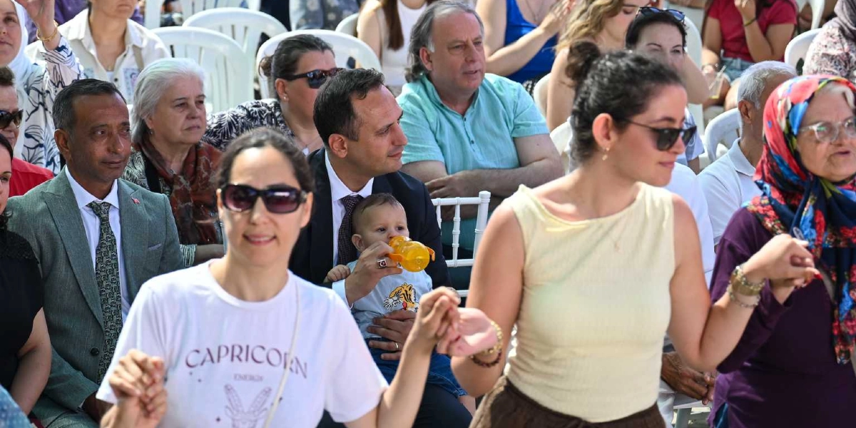 Açık havada düzenlenen bir etkinlikte, ön planda el ele tutuşarak halay çeken veya dans eden kadınlar, arka planda ise beyaz sandalyelerde oturarak onları izleyen çeşitli yaş gruplarından insanlar yer alıyor. Arka planda takım elbiseli bir adamın kucağında bebek tuttuğu, diğer katılımcıların ise günlük kıyafetlerle etkinliği takip ettiği görülüyor.