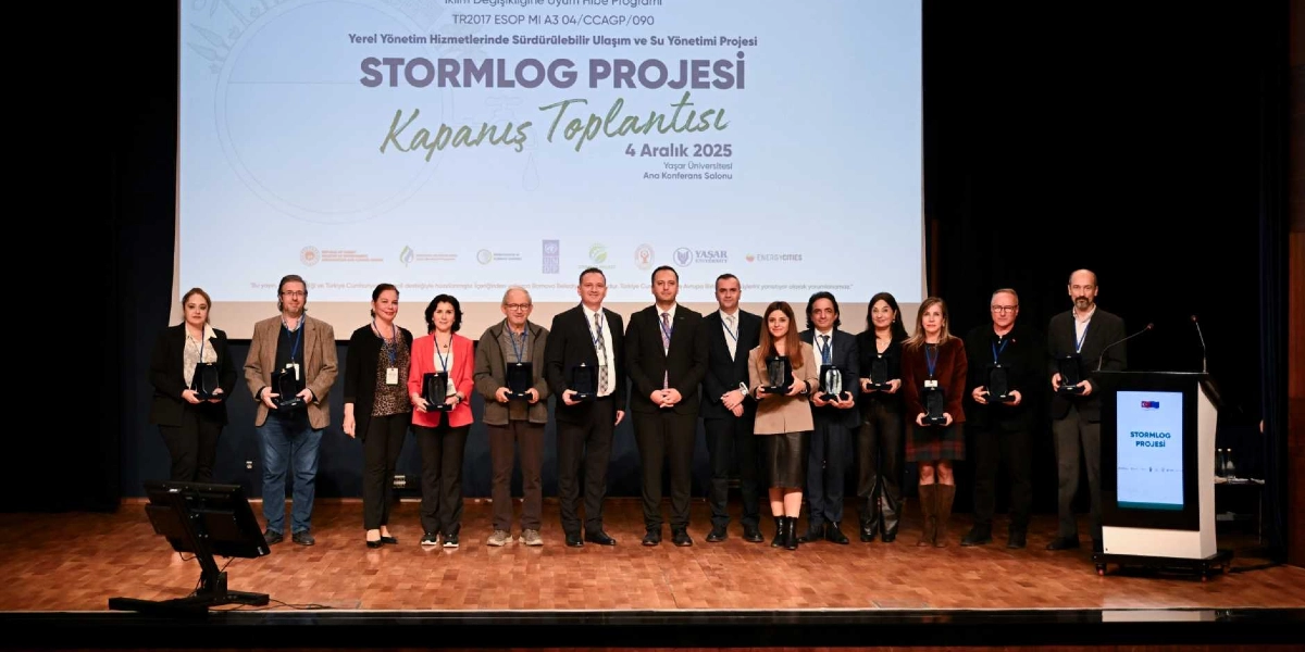Yaşar Üniversitesi Ana Konferans Salonu'nda düzenlenen STORMLOG Projesi Kapanış Toplantısı'nda, ellerinde plaketlerle yan yana duran katılımcı grup. Arka planda, projenin adının, tarihinin ve destekleyen kurumların logolarının yer aldığı büyük bir ekran bulunuyor. Sağ tarafta üzerinde STORMLOG PROJESİ yazılı bir kürsü yer alıyor.