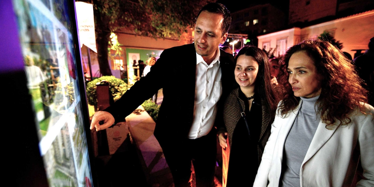 Gece dış mekanda, siyah takım elbiseli bir erkek, yanındaki iki kadına dijital bir ekran üzerindeki görselleri işaret ederek bir şeyler anlatıyor. Ekranın üzerinde Bornova'nın tarihsel mirasına dair olduğu anlaşılan görsel içerikler yer alıyor. Üç kişi de dikkatle ekrana bakıyor, arka planda ise ağaçlar ve aydınlatılmış bir çevre görülüyor.