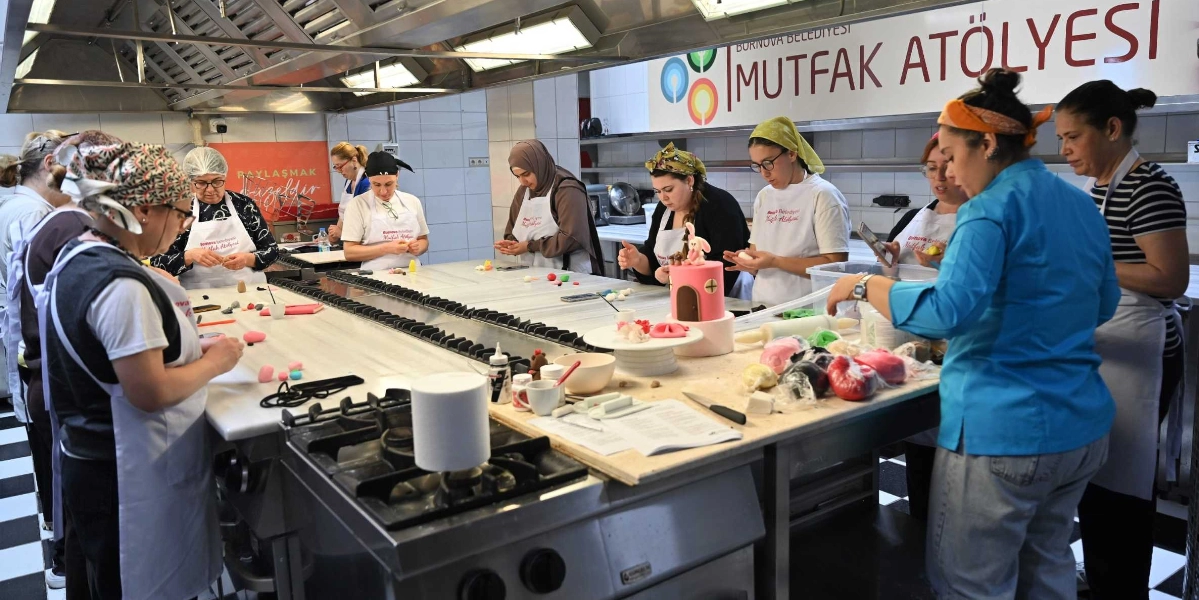 Bornova Belediyesi Mutfak Atölyesi'nde önlük giymiş bir grup kadın, uzun bir mutfak tezgahı etrafında toplanmış şeker hamuruyla pasta süsleme çalışması yapıyor. Katılımcılar önlerindeki renkli hamurları şekillendirirken, tezgahın üzerinde tamamlanmış küçük bir pasta figürü ve çeşitli mutfak gereçleri bulunuyor. Arka planda 