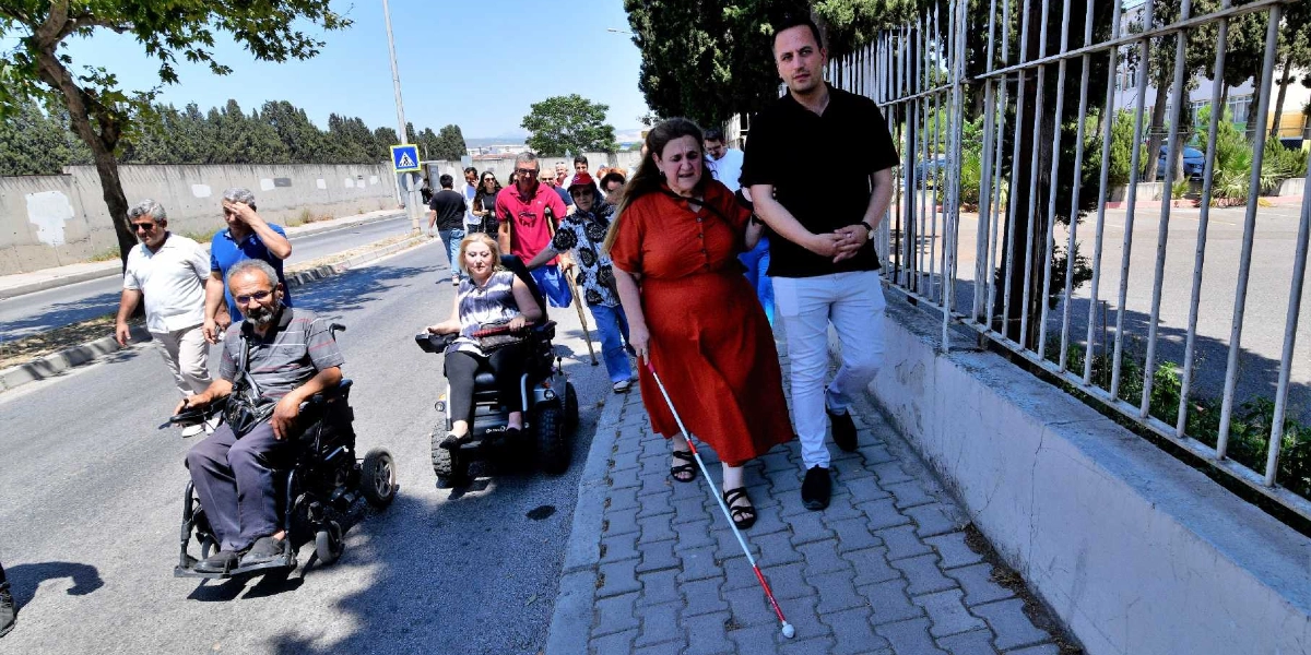 Güneşli bir günde kaldırımda ve yol kenarında yürüyen bir grup insan. Ön planda, beyaz baston kullanan turuncu elbiseli bir kadın, ona eşlik eden siyah tişörtlü bir erkekle birlikte kaldırımda yürüyor. Hemen yanlarında, yolda akülü tekerlekli sandalyede oturan bir kadın ve bir erkek bulunuyor. Grubun arkasında ise yürüyerek ilerleyen başka katılımcılar yer alıyor.