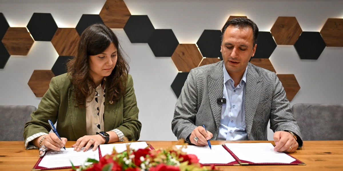 Modern bir ofis ortamında, altıgen ahşap panellerle süslü bir duvarın önünde, ahşap bir masa başında oturan bir kadın ve bir erkek resmi bir belgeyi imzalıyor. Kadın yeşil bir ceket, erkek ise gri ekose bir ceket giyiyor. Masanın ön kısmında kırmızı çiçeklerden oluşan bir aranjman bulunuyor.