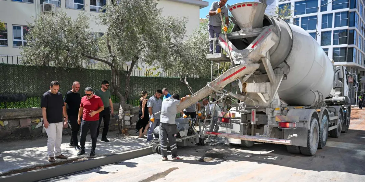 Şehir merkezinde bir sokakta, beton mikserinden dökülen harçla otomatik bordür taşı döşeme makinesini kullanan işçiler ve onları izleyen bir grup yetkili.