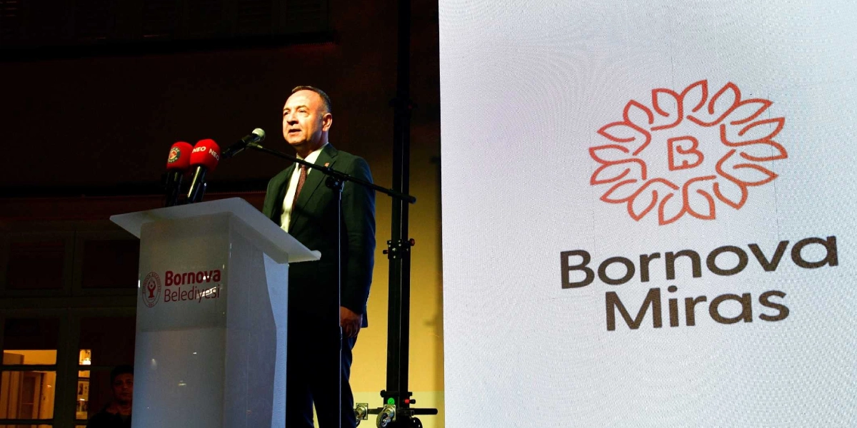 Koyu renk takım elbiseli bir erkek, üzerinde Bornova Belediyesi logosu bulunan bir kürsüde mikrofon başında konuşma yapıyor. Yanındaki büyük beyaz ekranda ise stilize edilmiş bir çiçek figürü ve altında Bornova Miras yazısı yer alıyor.