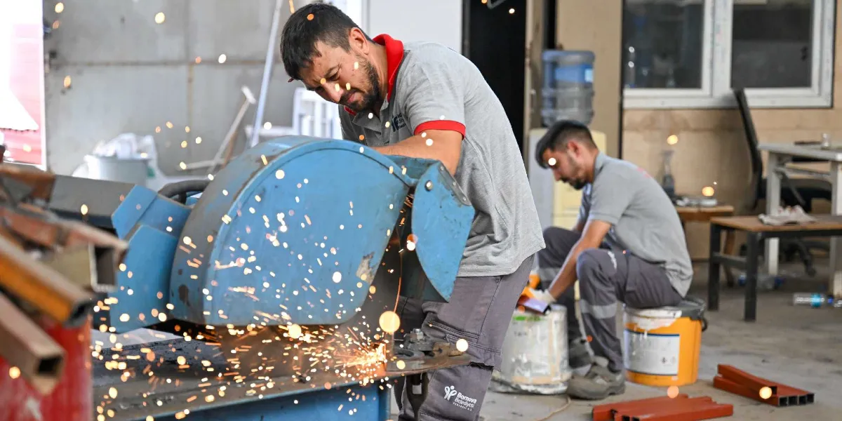 Bornova Belediyesi atölyesinde, üzerinde Bornova Belediyesi logosu bulunan gri iş kıyafetli bir çalışan, mavi bir metal kesme makinesiyle demir profil kesiyor. Kesim sırasında etrafa kıvılcımlar saçılıyor. Arka planda ise başka bir çalışan oturarak benzer metal parçaları üzerinde çalışmaya devam ediyor.