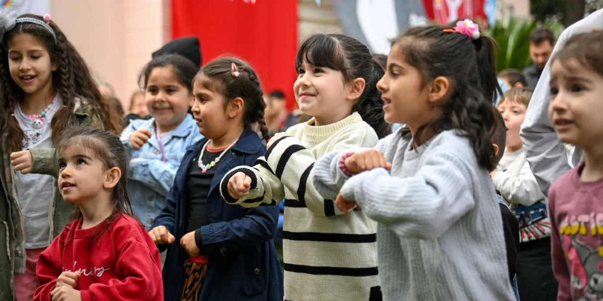 Açık havada düzenlenen bir etkinlikte, neşeyle dans eden ve şarkı söyleyen bir grup çocuk. Ön planda kırmızı sweatshirt giyen küçük bir kız çocuğu, yanında ise çizgili kazaklı ve gri sweatshirtlü diğer çocuklar yer alıyor. Arka planda kırmızı bayrakların asılı olduğu, şenlik havasının hakim olduğu bir ortam görülüyor.