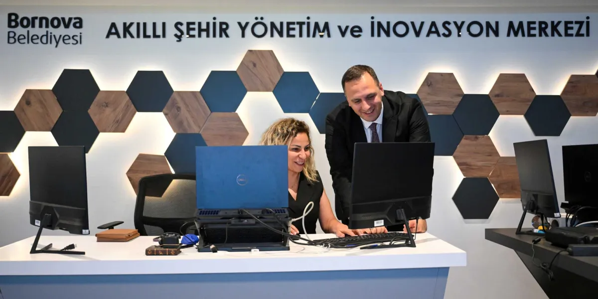 Bornova Belediyesi Akıllı Şehir Yönetim ve İnovasyon Merkezi'nde, beyaz bir çalışma masasında oturan bir kadın ve yanında ayakta duran bir erkek, bilgisayar ekranına bakarak gülümseyerek çalışıyor. Arka planda altıgen ahşap ve koyu renkli panellerden oluşan modern bir duvar dekorasyonu bulunuyor.