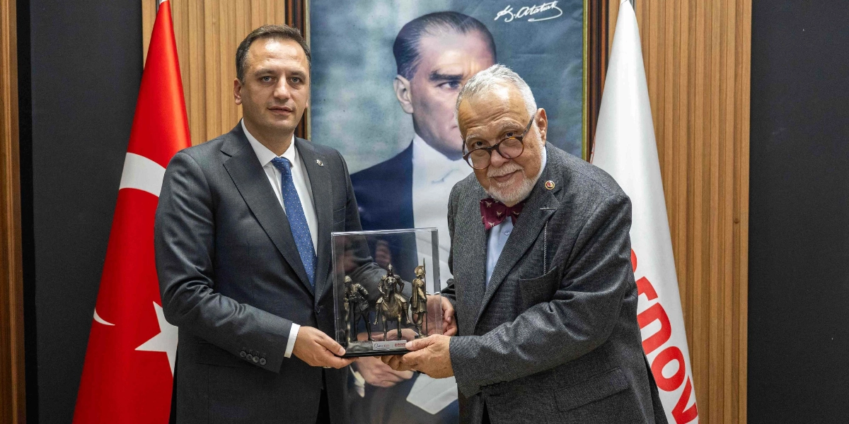 Bornova Belediye Başkanı Mustafa İduğ ve İlber Ortaylı, arkalarında Mustafa Kemal Atatürk portresi ve Türk bayrağı bulunan bir odada, üzerinde tarihi figürlerin yer aldığı cam muhafazalı bir heykeli birlikte tutuyorlar.