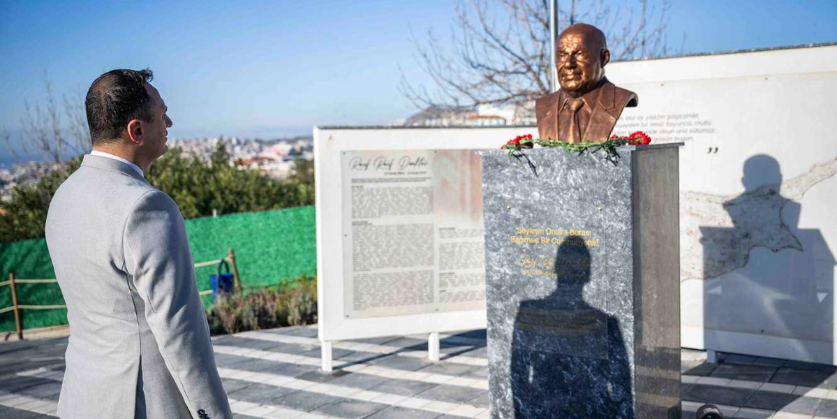 Açık renk ceket giymiş bir erkek, Kuzey Kıbrıs Türk Cumhuriyeti'nin kurucu Cumhurbaşkanı Rauf Denktaş'ın büstüne saygı duruşunda bulunuyor. Mermer kaide üzerinde yer alan büstün önünde kırmızı karanfiller bulunuyor ve kaidenin üzerinde 