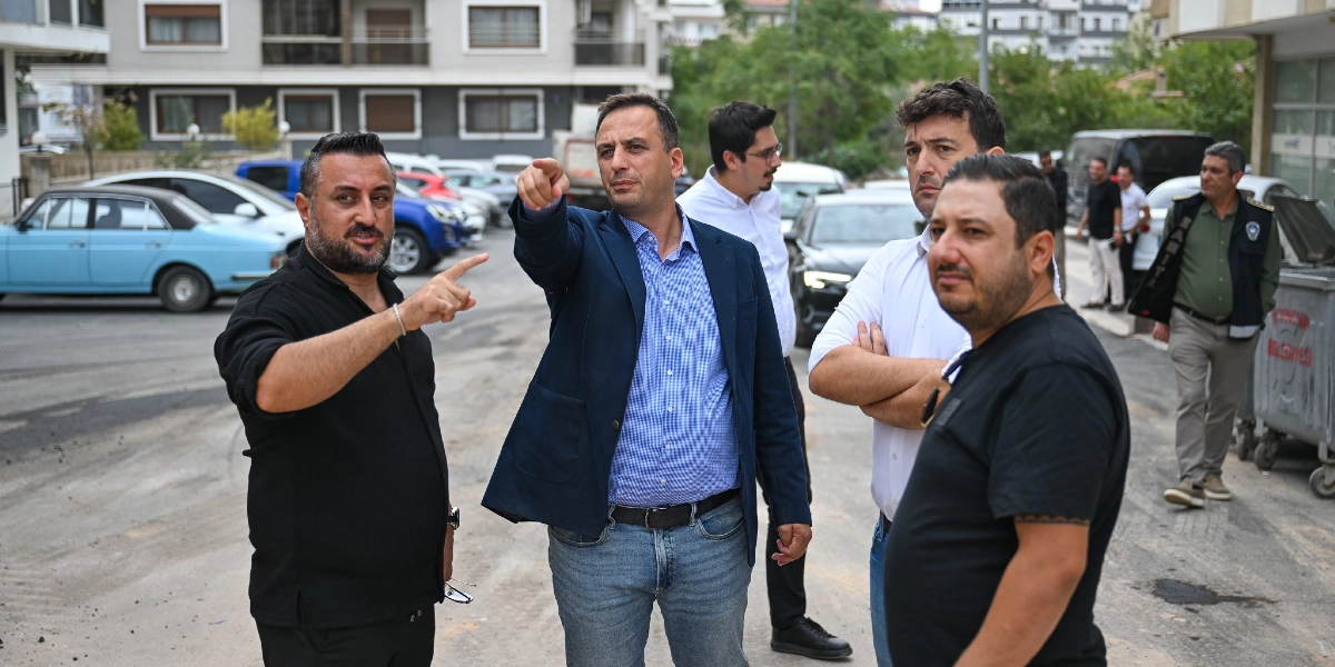 Şehir içinde bir sokakta bir araya gelmiş beş kişilik bir grup, çevre düzenlemesi veya kentsel dönüşüm çalışmalarıyla ilgili incelemelerde bulunuyor. Ön planda yer alan iki erkekten biri elini ileriye doğru uzatarak bir noktayı işaret ederken, yanındaki diğer kişi de benzer şekilde işaret ederek bir şeyler anlatıyor. Grubun geri kalanı onları dinliyor; arka planda ise park halindeki araçlar, apartmanlar ve üzerinde belediye yazısı bulunan büyük bir çöp konteyneri görülüyor.