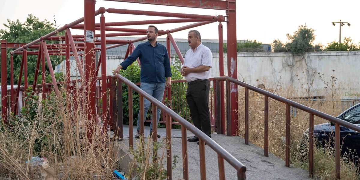 Kırmızı metal bir yaya üst geçidinin üzerinde duran iki erkek, çevreyi inceleyerek sohbet ediyor. Lacivert gömlekli ve kot pantolonlu kişi sol tarafta, beyaz kısa kollu gömlekli ve koyu renk pantolonlu kişi ise sağ tarafta yer alıyor. Arka planda beton bir duvar, kurumuş otlar ve kısmen görünen bir otomobil bulunuyor.