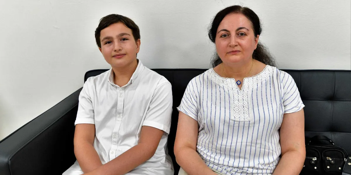 Beyaz duvar önünde siyah bir koltukta yan yana oturan, beyaz gömlekli genç bir erkek ve beyaz üzerine dikey çizgili bluz giyen orta yaşlı bir kadın kameraya bakıyor.