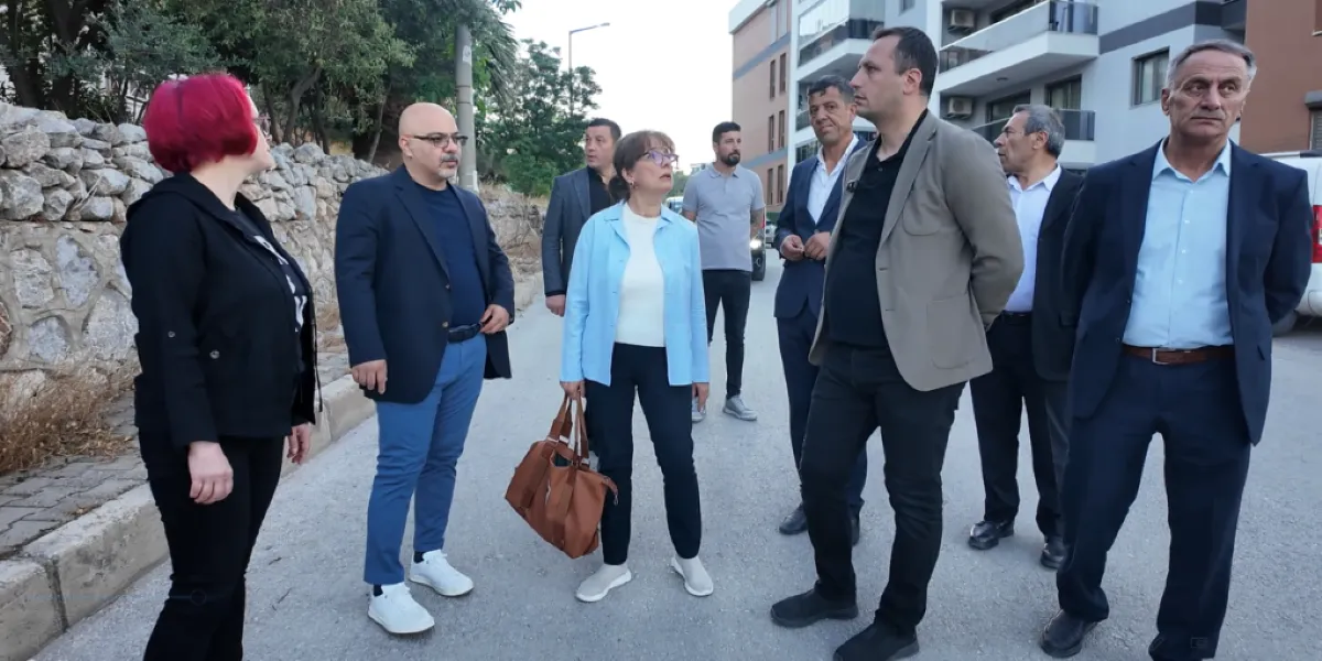 Bornova İzyuva'da bir sokakta bir araya gelmiş, aralarında yerel yöneticilerin de bulunduğu bir grup insan, çevre düzenlemesi veya yapılaşma üzerine incelemelerde bulunuyor. Taş duvarlı bir yol kenarında duran grupta, açık mavi ceketli ve elinde kahverengi bir çanta taşıyan bir kadın ile yanında takım elbiseli erkekler yer alıyor. Arka planda modern apartmanların bulunduğu sokakta, grup üyeleri dikkatle çevreyi gözlemliyor ve birbirleriyle iletişim kuruyor.
