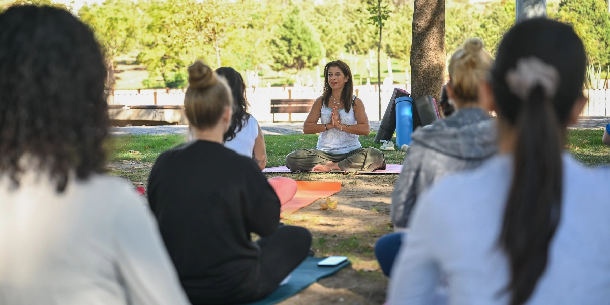 Açık havada, yeşil bir alanda yoga matları üzerinde bağdaş kurarak oturan bir grup kadın, eğitmen eşliğinde meditasyon yapıyor. Eğitmen, grubun önünde matının üzerinde oturmuş, ellerini göğüs hizasında birleştirmiş şekilde duruyor.