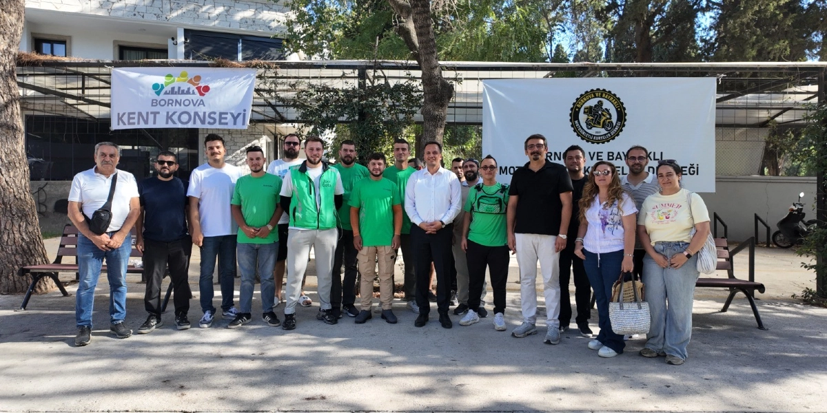 Açık havada, ağaçların önünde yan yana duran yaklaşık 20 kişilik bir grup moto kurye ve temsilci. Arka planda üzerinde Bornova Kent Konseyi yazılı bir pankart ile Bornova ve Bayraklı Motosikletli Kuryeler Derneği logosunun bulunduğu ikinci bir pankart yer alıyor. Grubun büyük çoğunluğu günlük kıyafetler giyerken, bazıları üzerinde kurye firmalarının logoları bulunan yeşil yelekler taşıyor. Grupta hem erkek hem de kadın katılımcılar bulunuyor; herkes kameraya bakarak gülümsüyor.