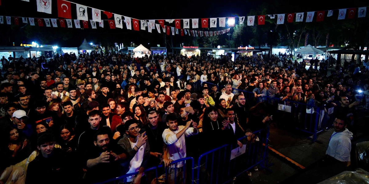 Gece vakti düzenlenen açık hava festivalinde, bariyerlerin önünde toplanmış büyük bir kalabalık sahneye doğru bakıyor. İnsanların çoğu gençlerden oluşuyor ve bazıları telefonlarıyla çekim yapıyor. Alanın üzerinde, Türk bayrakları ve üzerinde Atatürk silüeti bulunan flamaların birbirine bağlandığı bir süsleme dizisi uzanıyor. Arka planda ise ışıklandırılmış büfeler ve stantlar görülüyor.