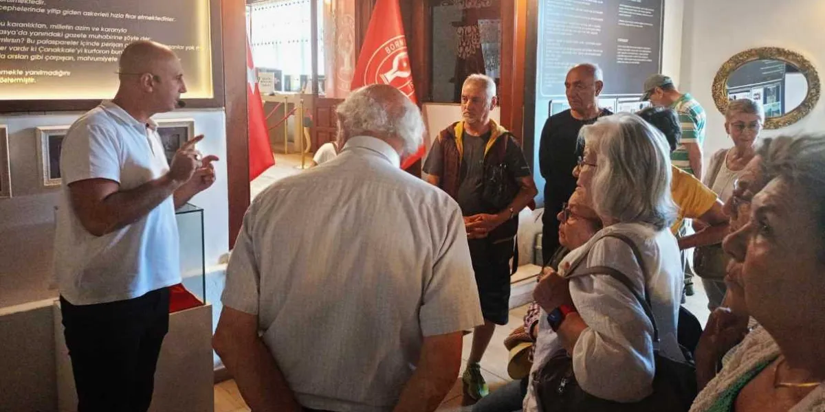 Tarihi bir mekanda veya müzede rehberlik yapan beyaz tişörtlü bir adam, etrafındaki katılımcı grubuna bilgi veriyor. Katılımcılar arasında ağırlıklı olarak orta yaş ve üzeri bireyler bulunuyor. Arka planda Bornova Belediyesi logosunun yer aldığı kırmızı bir bayrak, duvarlarda bilgilendirici metin panoları ve çerçeveli görseller yer alıyor. Mekan, tarihsel bir atmosfer yansıtan aydınlatmaya sahip.