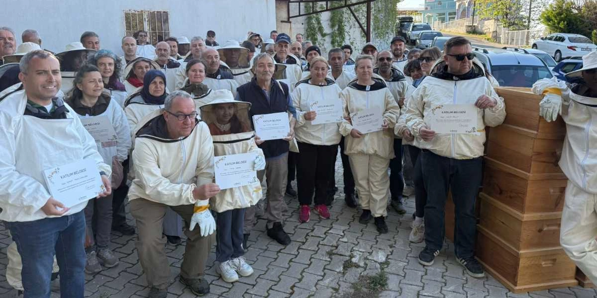 Arıcılık eğitimi almış, üzerlerinde koruyucu beyaz arıcı kıyafetleri bulunan kalabalık bir grup, ellerinde katılım belgeleriyle dış mekanda poz veriyor. Grubun sağ tarafında üst üste dizilmiş ahşap arı kovanları yer alıyor. Arka planda ise park halindeki araçlar ve ağaçlar görülüyor.