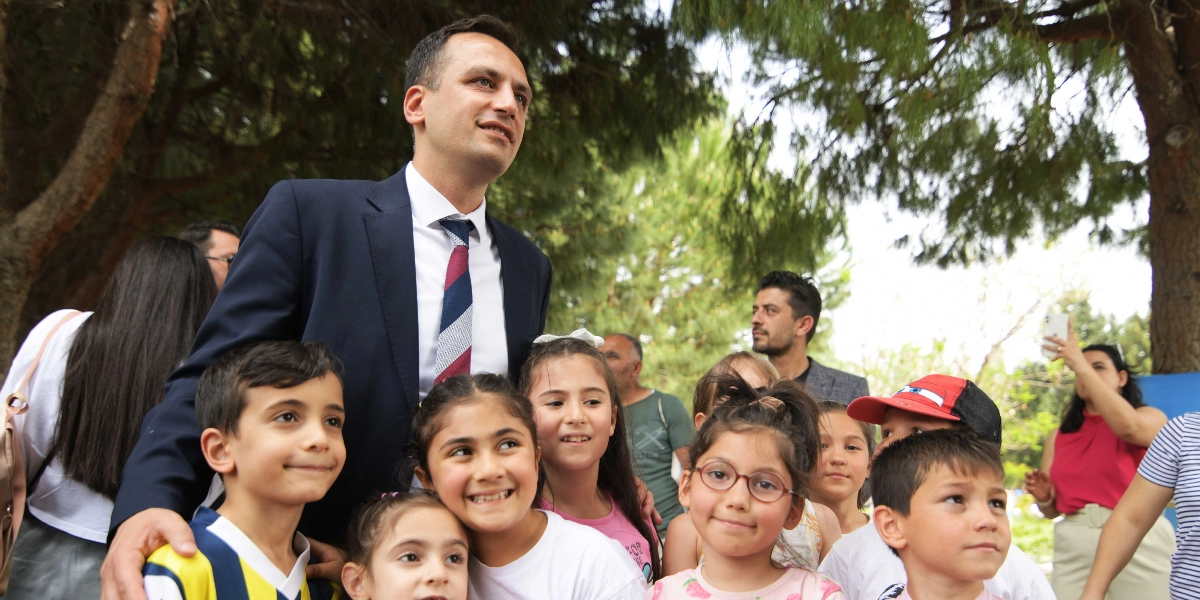 Lacivert takım elbiseli ve çizgili kravatlı bir erkek, park ortamında etrafını saran bir grup çocukla birlikte gülümsüyor. Çocuklar ön planda yer alırken, arka planda ağaçlar ve etkinliği izleyen diğer yetişkinler görülüyor.