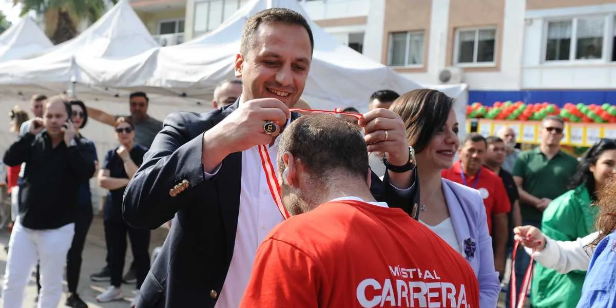 Koyu renk ceketli bir erkek, kırmızı tişörtlü bir katılımcının boynuna madalya takarken gülümsüyor. Yanlarında açık renk ceketli bir kadın ve arka planda beyaz çadırların önünde duran kalabalık bir grup yer alıyor. Kırmızı tişörtün üzerinde beyaz harflerle Mistral Carrera yazısı bulunuyor.