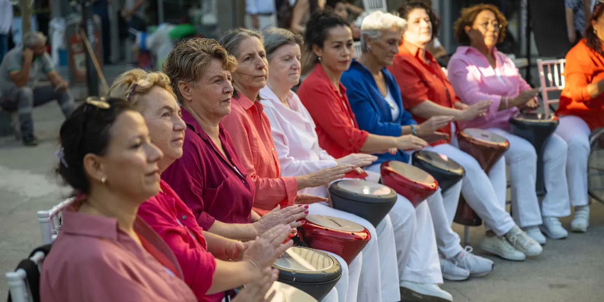 Yan yana dizilmiş sandalyelerde oturan bir grup kadın, önlerinde duran darbuka ve benzeri vurmalı çalgıları çalarak ritim tutuyor. Farklı renklerde kıyafetler giymiş olan kadınlar, odaklanmış bir şekilde müzik yaparken görülüyor. Arka planda ise etkinliği izleyen veya çevrede bulunan diğer insanlar seçiliyor.