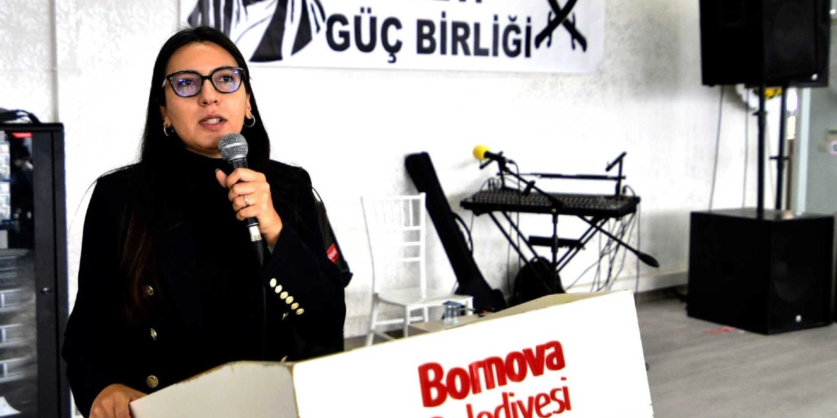 Siyah ceketli, gözlüklü bir kadın, üzerinde Bornova Belediyesi yazan bir kürsüde mikrofonla konuşma yapıyor. Arka planda üzerinde Alevi Güç Birliği yazan bir pankart, ses sistemi ekipmanları ve beyaz bir sandalye bulunuyor.