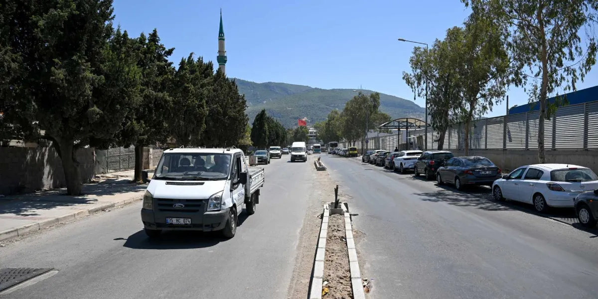 Güneşli bir günde, orta refüjü yenilenmekte olan Pınar Caddesi'nde trafik akışı devam ediyor. Yolun sol tarafında ağaçlar ve ileride bir cami minaresi, sağ tarafında ise park etmiş araçlar ve metal bir çit bulunuyor. Ön planda beyaz bir kamyonet cadde boyunca ilerliyor.
