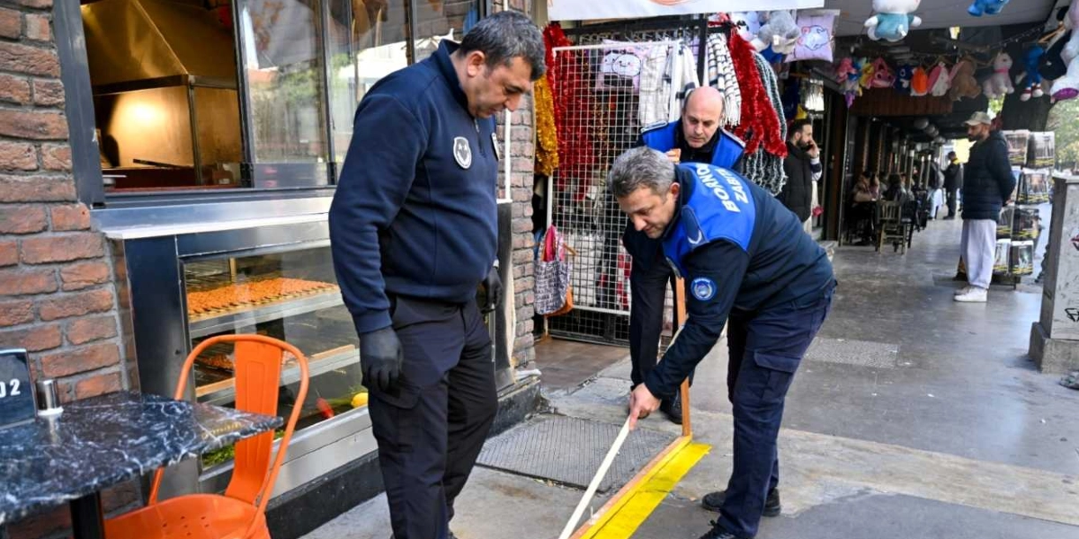 Bornova Belediyesi zabıta ekipleri, bir işletmenin önündeki kaldırımda sarı şerit kullanarak işgali belirlemek amacıyla ölçüm yapıyor. Üniformalı iki zabıta memuru eğilmiş şekilde yerdeki sarı çizgiyi kontrol ederken, bir diğer memur ise onları izliyor. Arka planda hediyelik eşya dükkanı ve kaldırımda yürüyen insanlar görülüyor.