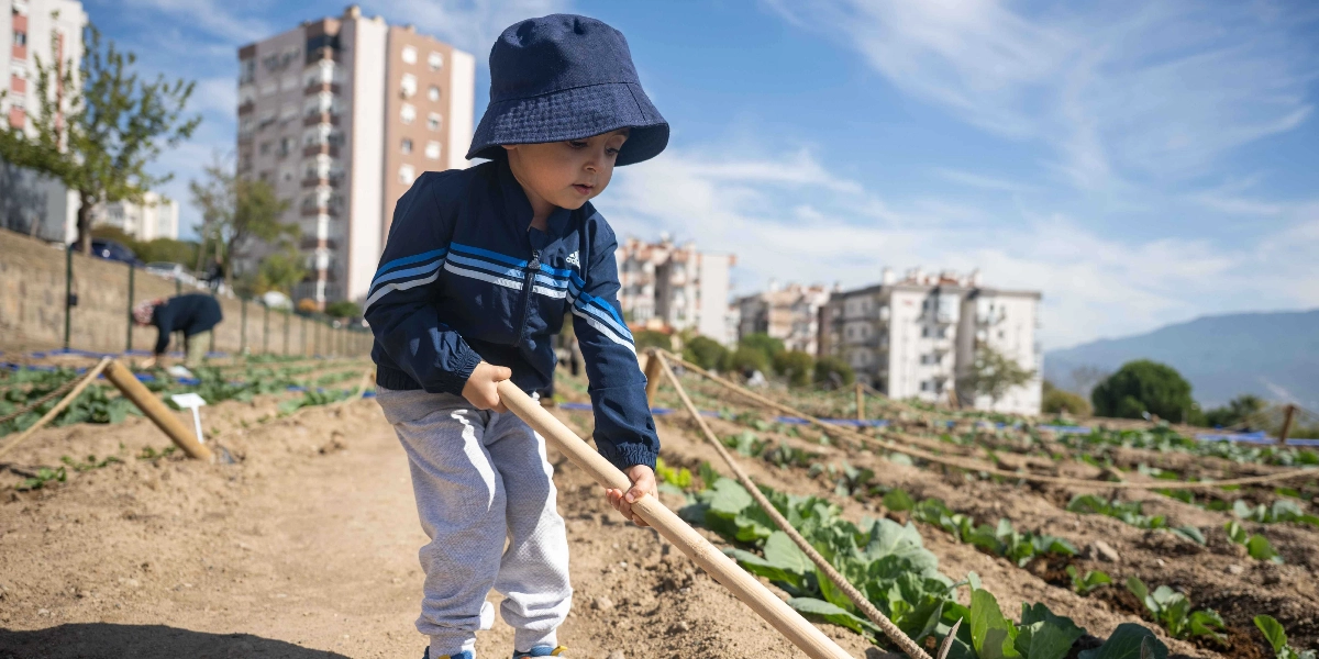 Lacivert şapkalı ve eşofmanlı küçük bir çocuk, şehir içindeki bir bostanda elindeki ahşap saplı çapa ile toprakla uğraşıyor. Arka planda çok katlı binalar ve güneşli bir gökyüzü görülürken, ileride toprakla ilgilenen başka bir kişi daha bulunuyor.