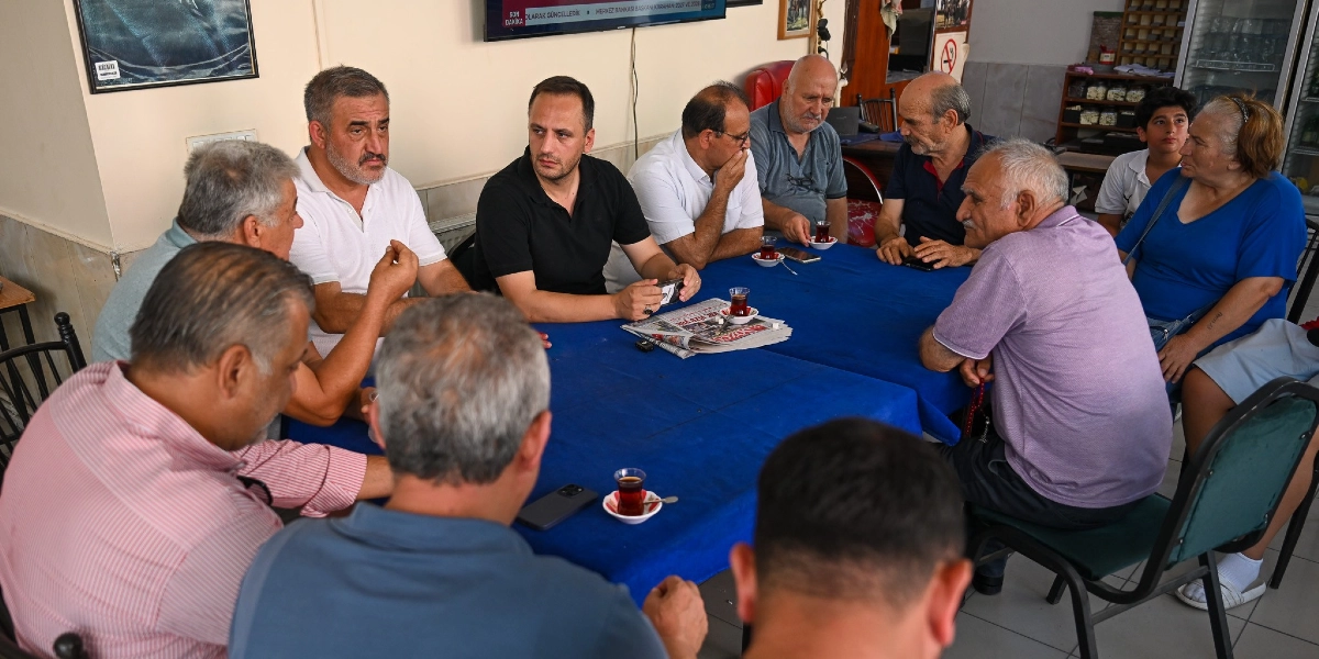 Mavi örtülü bir masanın etrafında toplanmış bir grup vatandaşla sohbet eden Başkan Eşki, masanın orta kısmında siyah tişörtüyle oturuyor. Çevresindeki masada oturan kişiler çaylarını yudumlarken, masanın üzerinde bir gazete ve akıllı telefonlar bulunuyor. İç mekanda gerçekleşen bu buluşmada, katılımcılar dikkatle dinliyor ve karşılıklı fikir alışverişinde bulunuyor.