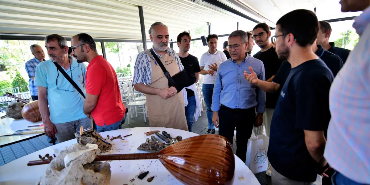 Açık havada, üstü kapalı bir alanda toplanmış bir grup insan, masanın üzerinde duran el yapımı bir bağlamayı ve yanında bulunan ahşap oyma aletlerini inceliyor. Grubun ortasında, üzerinde bej rengi bir önlük olan ve kolu askıda bulunan bir zanaatkar, çevresindekilere enstrüman yapımı hakkında bilgi veriyor. Masanın üzerinde tamamlanmış bir bağlamanın yanı sıra, ağaç parçaları ve çeşitli kesici aletler yer alıyor. Etraftaki katılımcılar ilgiyle süreci takip ederken, arka planda beyaz sandalyeler ve yeşil bir bahçe manzarası görünüyor.