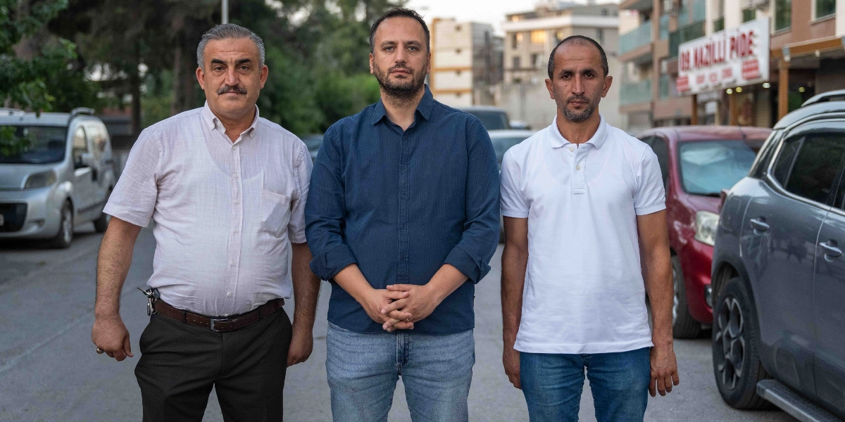 Sokak ortasında yan yana duran üç erkek; soldaki kişi açık renkli kısa kollu gömlek ve koyu renkli pantolon, ortadaki kişi lacivert uzun kollu gömlek ve kot pantolon, sağdaki kişi ise beyaz polo yaka tişört ve kot pantolon giymiş durumda. Arka planda park halindeki araçlar ve sağ tarafta 