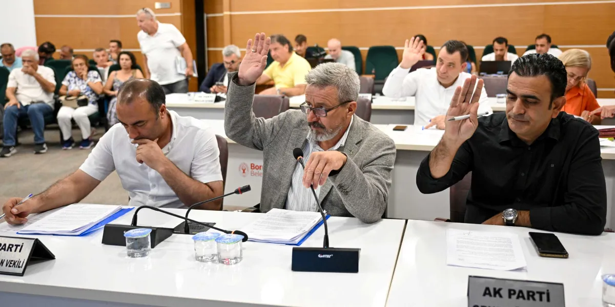 Bornova Belediyesi meclis toplantısında, masada oturan katılımcılardan bazıları oylama yapmak için ellerini kaldırıyor. Ön planda, üzerinde mikrofonlar, belgeler ve su bardakları bulunan beyaz bir masada oturan üç erkekten ortadaki gri ceketli ve gözlüklü kişi ile sağdaki siyah gömlekli kişi elini kaldırmış durumda. Soldaki beyaz gömlekli kişi ise önündeki belgelere bakıyor. Arka planda toplantıya katılan diğer kişiler sıralar halinde oturuyor ve masaların üzerinde 