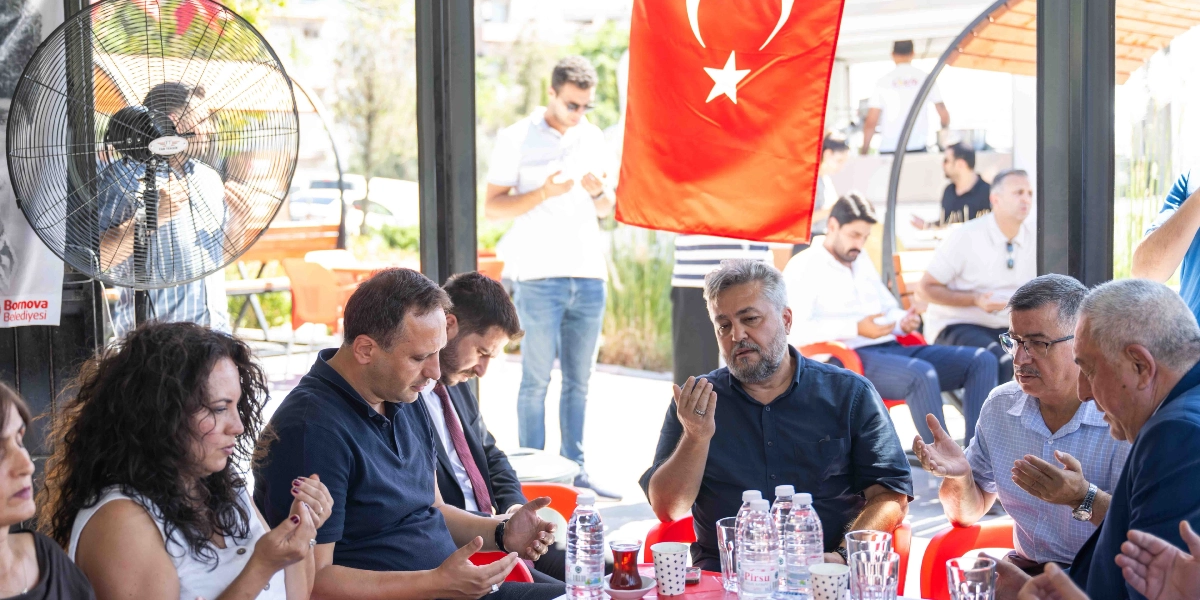 Açık havada, üzerinde Türk bayrağı asılı bir mekanda bir araya gelen bir grup insan, ellerini açarak dua ediyor. Masada oturan kadın ve erkekler hüzünlü bir ifadeyle dua ederken, arka planda ayakta duranlar da onlara eşlik ediyor. Masanın üzerinde su şişeleri ve çay bardakları bulunuyor, sol tarafta ise Bornova Belediyesi logosunun yer aldığı bir afiş ve büyük bir vantilatör görülüyor.