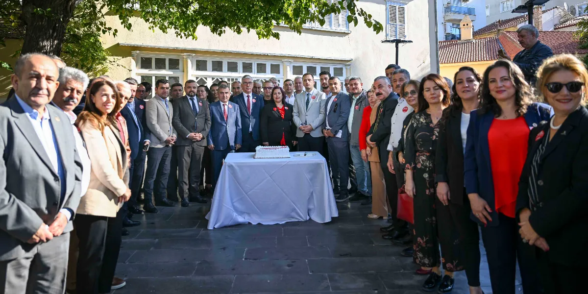Açık havada, taş zeminli bir avluda toplanmış, aralarında Bornova Belediye Başkanı Mustafa İduğ'un da bulunduğu kalabalık bir grup insan, ortadaki beyaz örtülü masanın üzerinde duran pastanın etrafında duruyor. Resmi kıyafetli kadın ve erkeklerden oluşan grup, Muhtarlar Akademisi'nin açılışını kutlamak üzere bir arada poz veriyor.