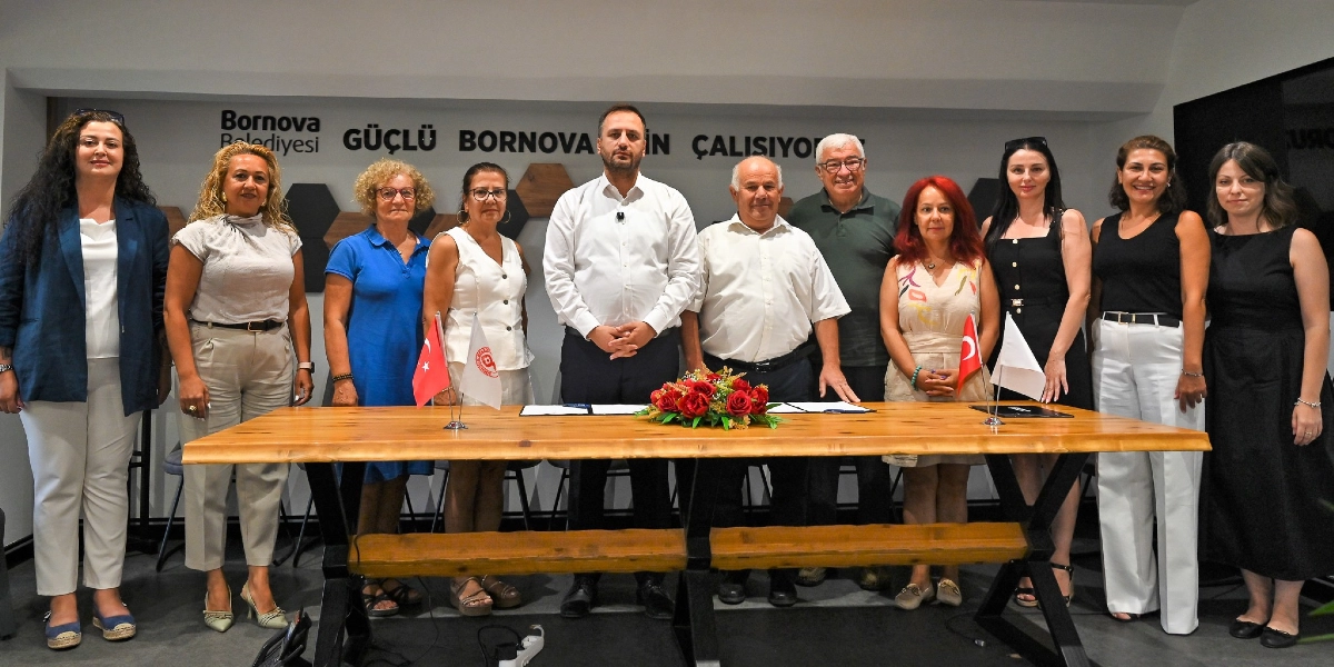 Bornova Belediyesi'nde gerçekleşen işbirliği protokolü imza töreninde, ahşap bir masanın arkasında yan yana duran on kişilik bir grup yer alıyor. Grubun merkezinde, beyaz gömlekli bir erkek ve yanında açık renkli gömlekli bir erkek bulunuyor. Masanın üzerinde kırmızı çiçeklerden oluşan bir aranjman ve iki küçük bayrak yer alıyor. Arka planda duvarda 