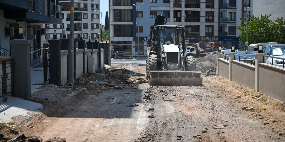 Bornova'da bir sokakta devam eden yol yenileme çalışmaları kapsamında, toprak ve çakıl zemin üzerinde ilerleyen bir iş makinesi görülüyor. Çevrede modern apartmanlar, dükkanlar ve park halindeki araçlar yer alıyor.