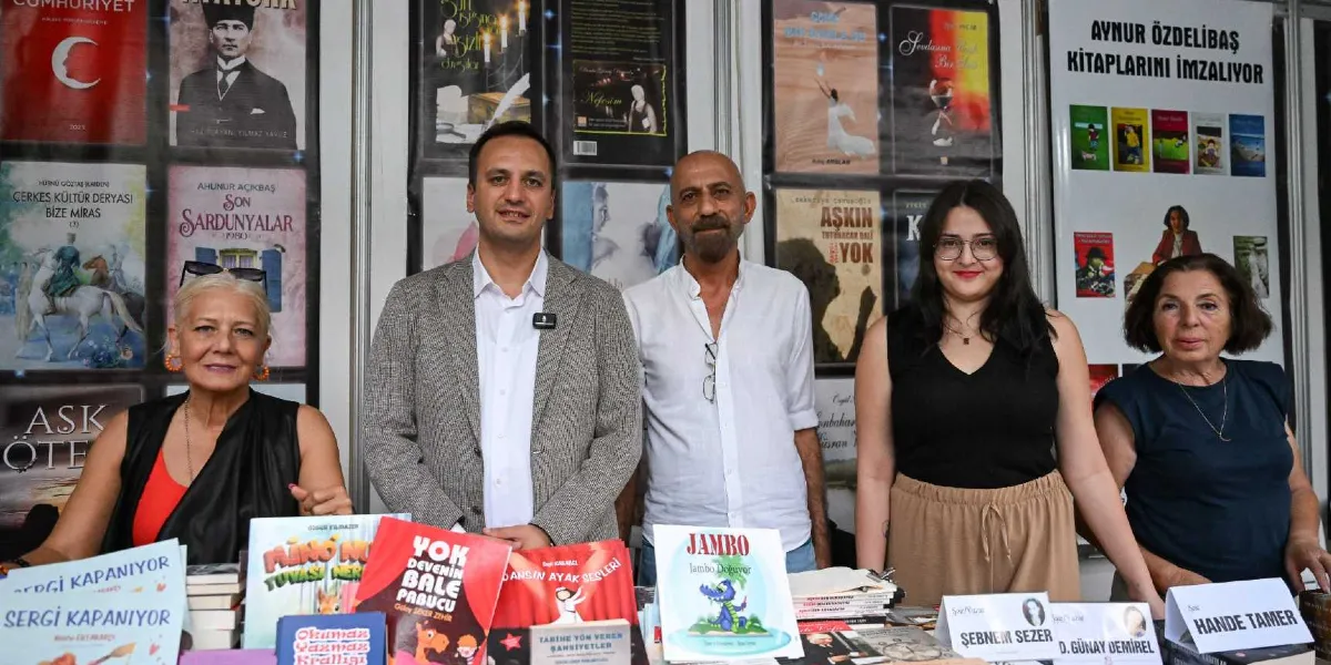 Bir kitap fuarı veya imza gününde, arka planda çeşitli kitap kapaklarının sergilendiği bir stant önünde duran beş kişi. Soldan sağa doğru; siyah yelekli bir kadın, gri ceketli bir erkek, beyaz gömlekli sakallı bir erkek, siyah kolsuz bluz giyen gözlüklü bir kadın ve lacivert bluzlu bir kadın yan yana duruyor. Masanın üzerinde 