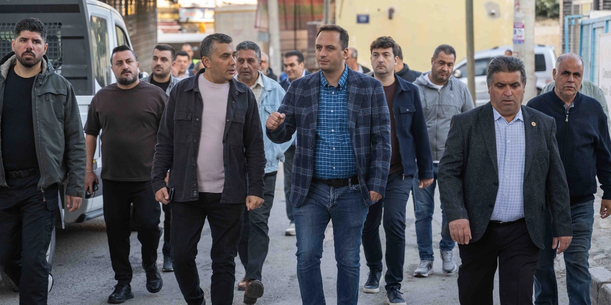 Bornova'da bir sokakta yürüyen, aralarında Bornova Belediye Başkanı Ömer Eşki'nin de bulunduğu, günlük kıyafetler giymiş bir grup erkek. Ömer Eşki, kareli ceketi ve kot pantolonuyla grubun önünde yer alarak konuşma yapıyor, yanındaki ve arkasındaki diğer kişiler ise onu dinleyerek birlikte ilerliyor.