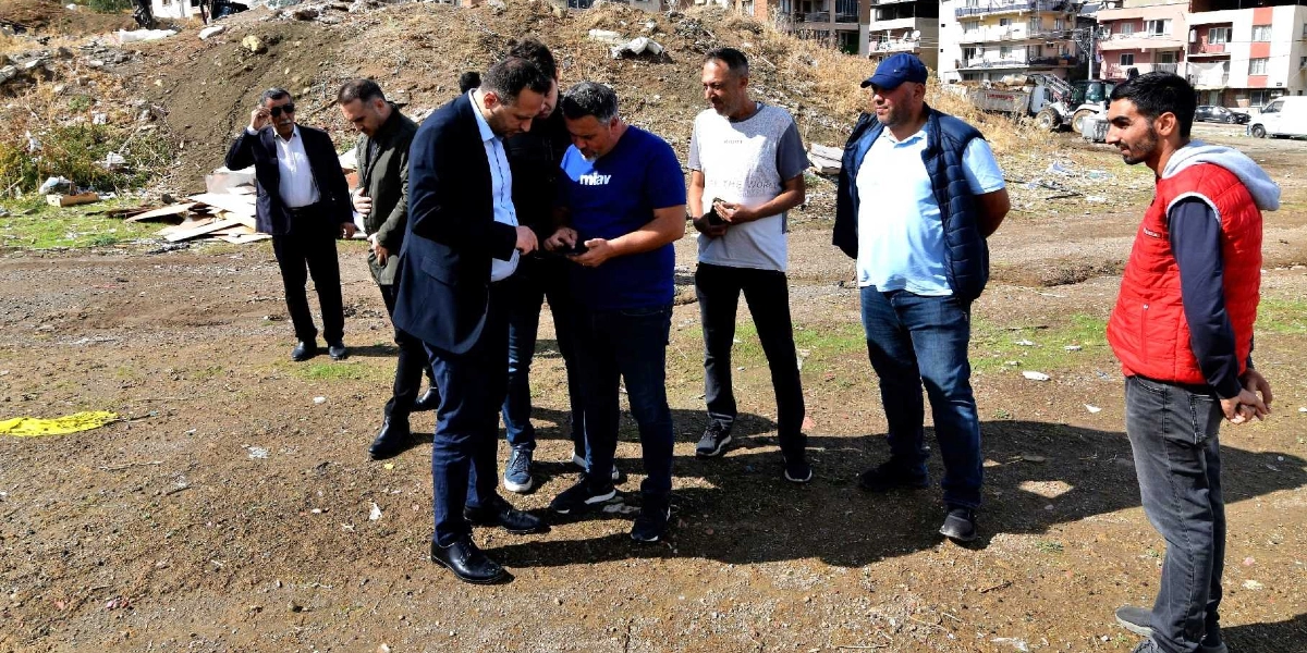Açık havada, toprak bir arazide bir araya gelmiş yedi kişilik bir grup, bir cep telefonunun ekranına bakarak inceleme yapıyor. Gruptakilerden biri lacivert takım elbiseli, diğeri üzerinde 