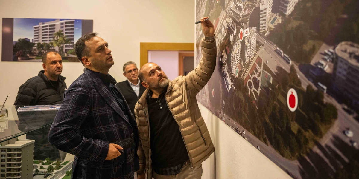 Ofis ortamında, duvarda asılı büyük bir şehir planı haritasını inceleyen üç erkek. Öndeki bej şişme montlu ve sakallı kişi, elindeki kalemle harita üzerindeki bir noktayı işaret ederek yanındaki koyu renk ekose ceketli kişiye bir şeyler anlatıyor. Arkada ise gözlüklü bir başka kişi onları izliyor. Harita üzerinde kırmızı nar simgeleri ve 