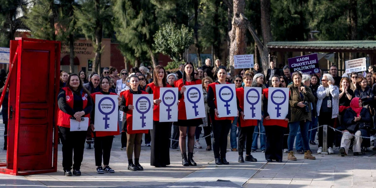 Açık havada, ağaçlık bir alanda düzenlenen 8 Mart etkinliğinde, kırmızı yelekler giymiş bir grup kadın yan yana dizilmiş, üzerinde kadın sembolü ve çeşitli sloganların yazılı olduğu beyaz dövizler tutuyor. Dövizlerin üzerinde sırasıyla 